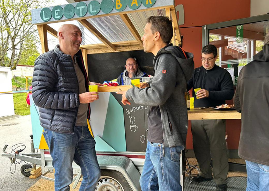 En extérieur, quatre hommes se tiennent de part et d'autres d'un comptoir au dessus duquel on peut lire LeTL bar