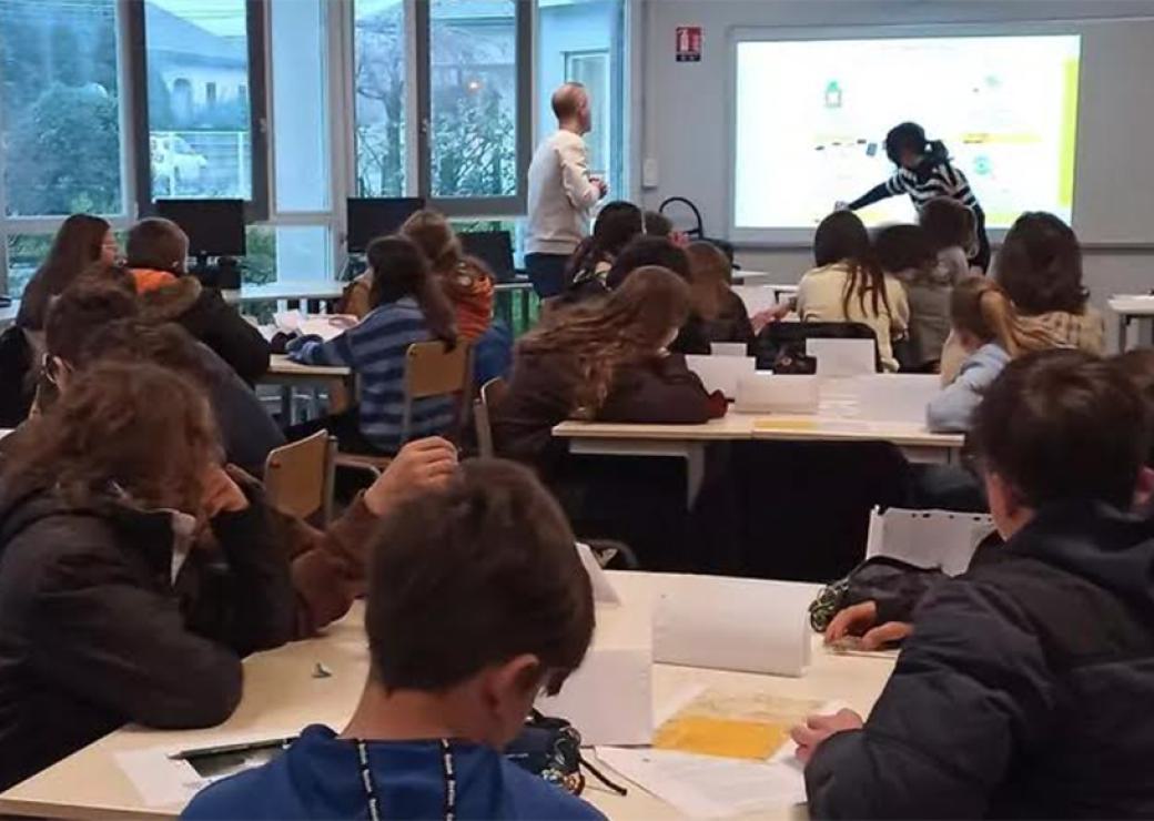 Dans une salle de classe des enfants regardent deux adultes qui sont devant un écran