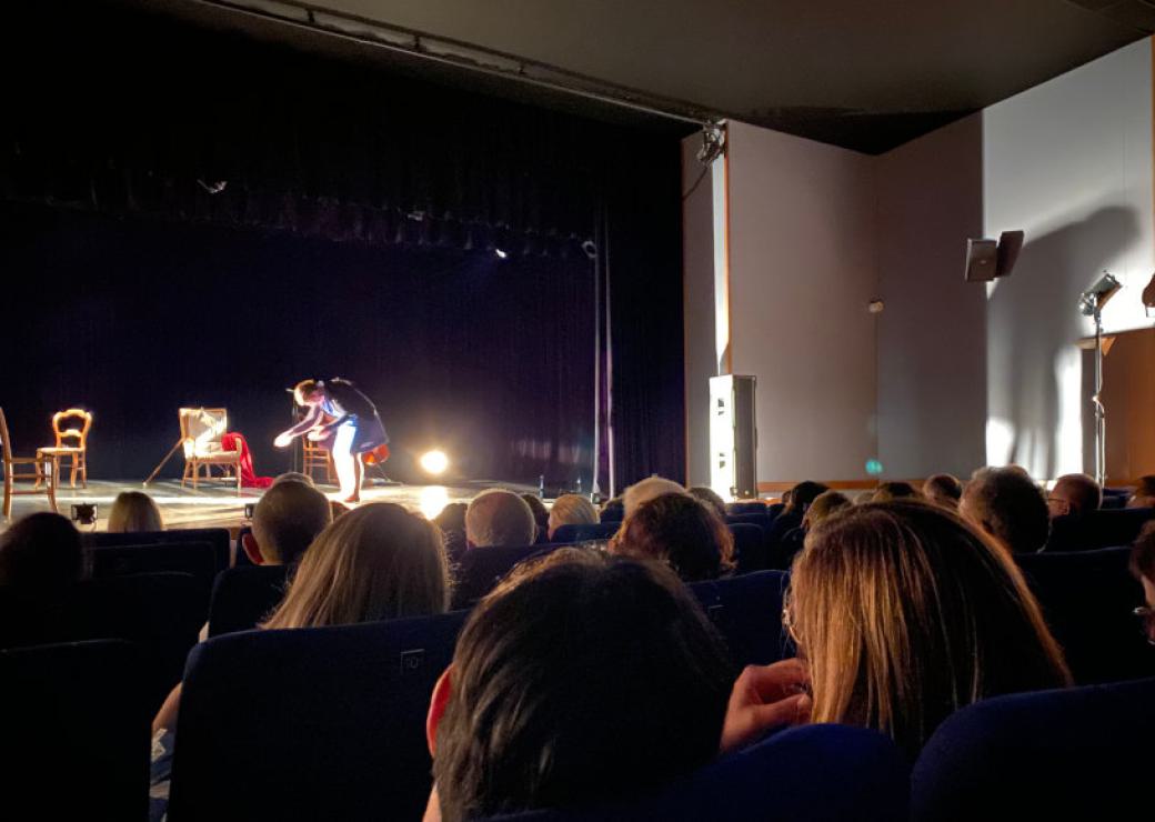 Dans une salle de spectacle, sur scène, une vieille femme très courbée