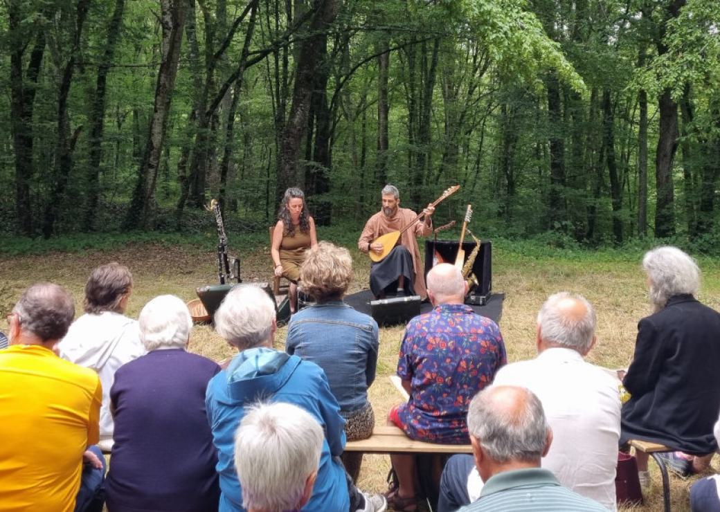 Un public assis sur des bancs dans une forêt, regardent deux musiciens qui leur font face