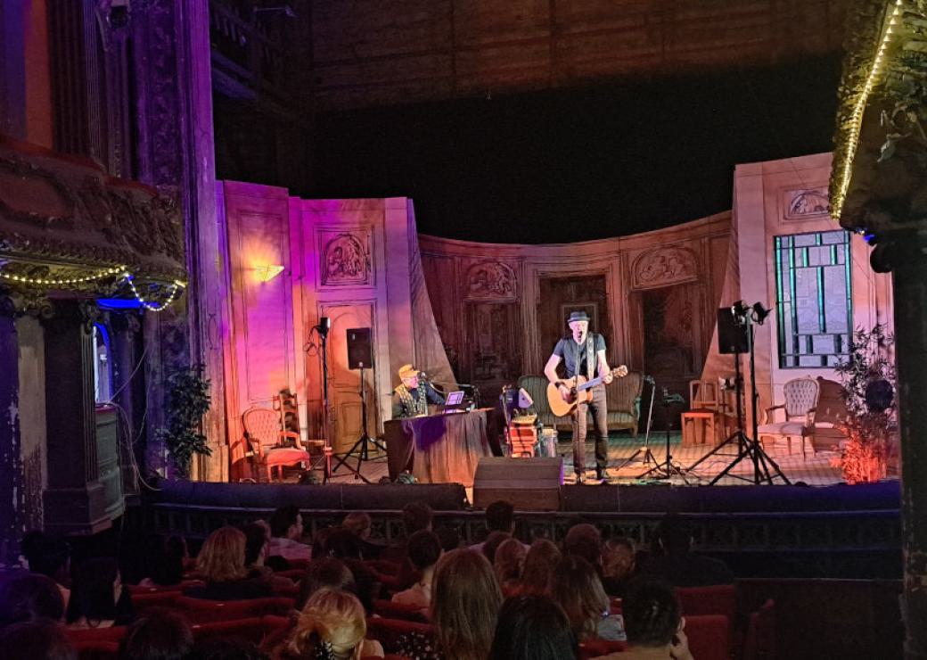 Vue depuis la salle de deux personnes sur une scène de théâtre, avec les décors et la lumière d'un spectacle