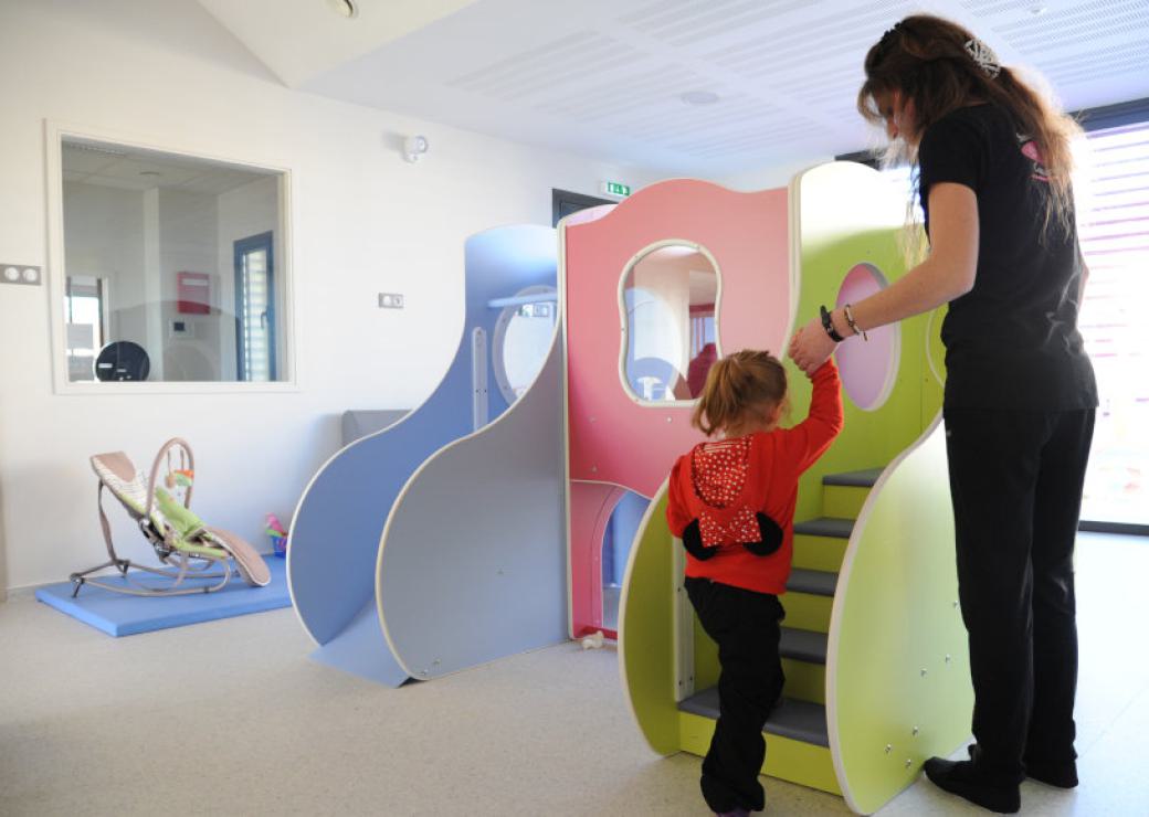 Une jeune femme aide une petite fille à monter les escaliers d'une structure de jeux, dans une salle aux murs blancs