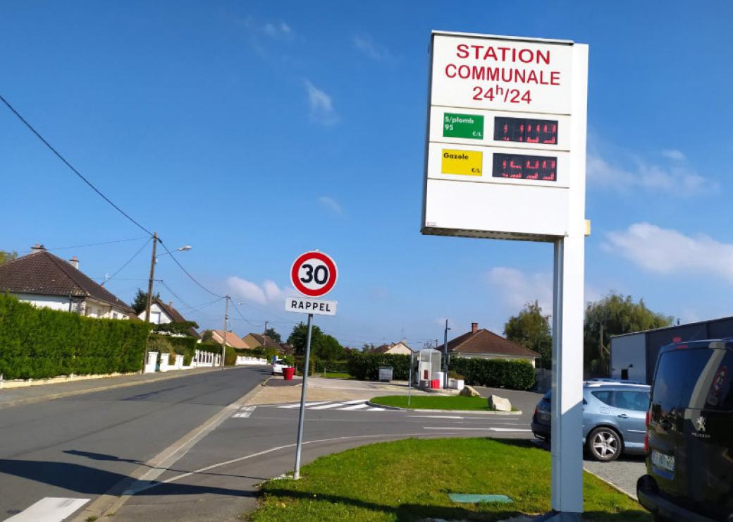 Sur le bord de la route, un panneau en hauteur indique les prix du carburant, sous l'enseigne Station service communale