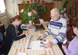 Dans le salon d'une maison, un homme et une femme âgés exposent sur la table des photos et des coupures de presse. Face à eux, une jeune femme prend des notes.