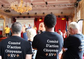 Trois personnes regardent en direction d'une scène installée dans une grande salle. Sur le dos de leur T-shirt, on peut lire "Comité d'action citoyenne XV"